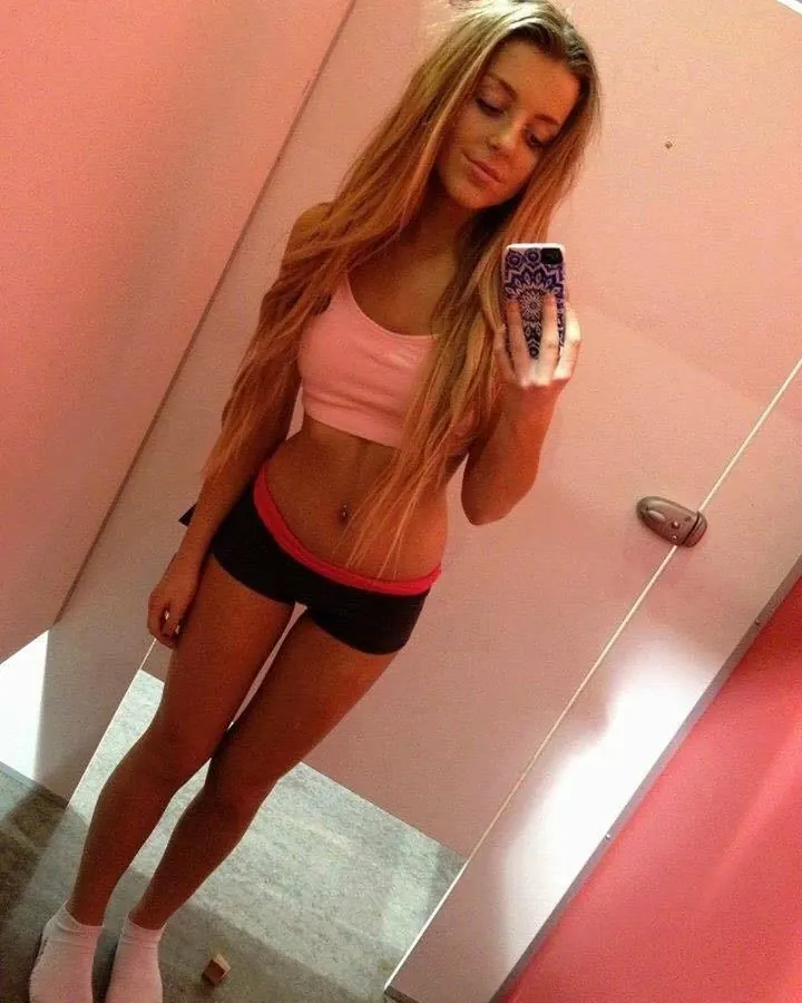Jeune femme en haut rose et short noir, prenant un selfie dans une salle rose