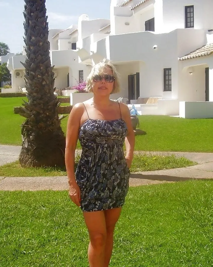Femme blonde en robe imprimée, debout sur une pelouse verte devant des bâtiments blancs