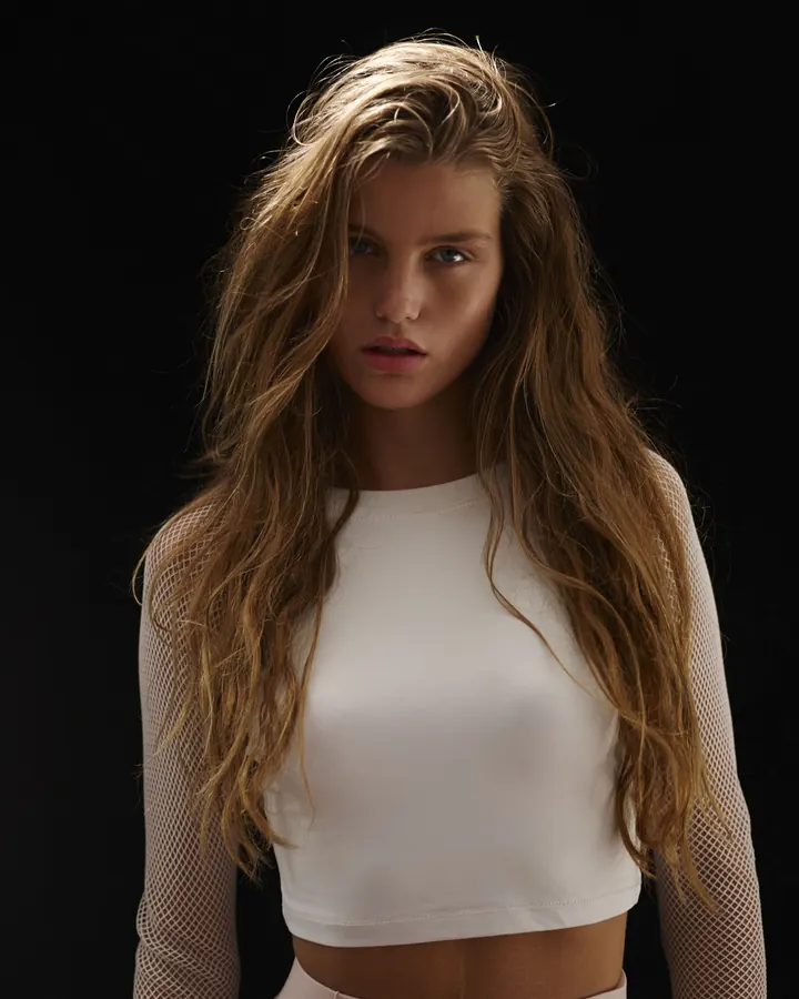 Femme aux cheveux bruns foncés portant un haut noir, posant de face avec un regard direct, sur fond blanc.