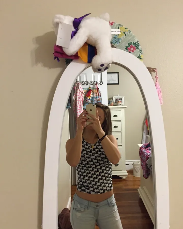 Jeune femme aux cheveux châtains, prenant un selfie devant un grand miroir, portant un haut imprimé noir et blanc et un jean déchiré.