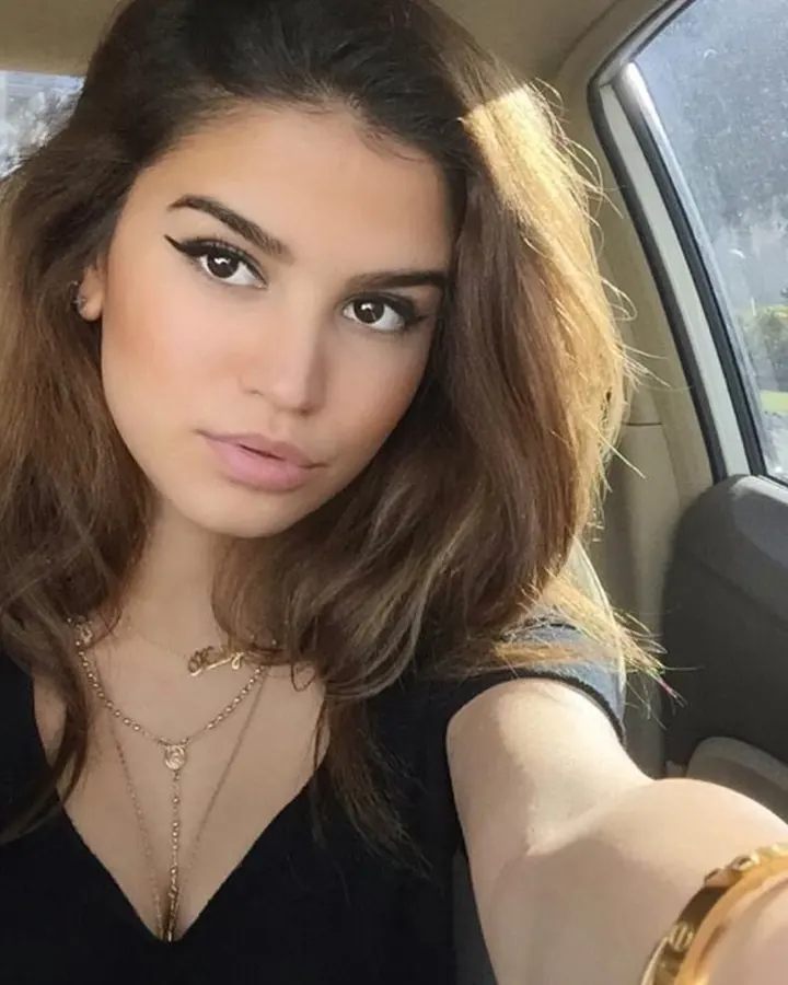 Femme aux cheveux bruns, assise dans une voiture, regardant la caméra, portant un haut noir et des bijoux dorés.