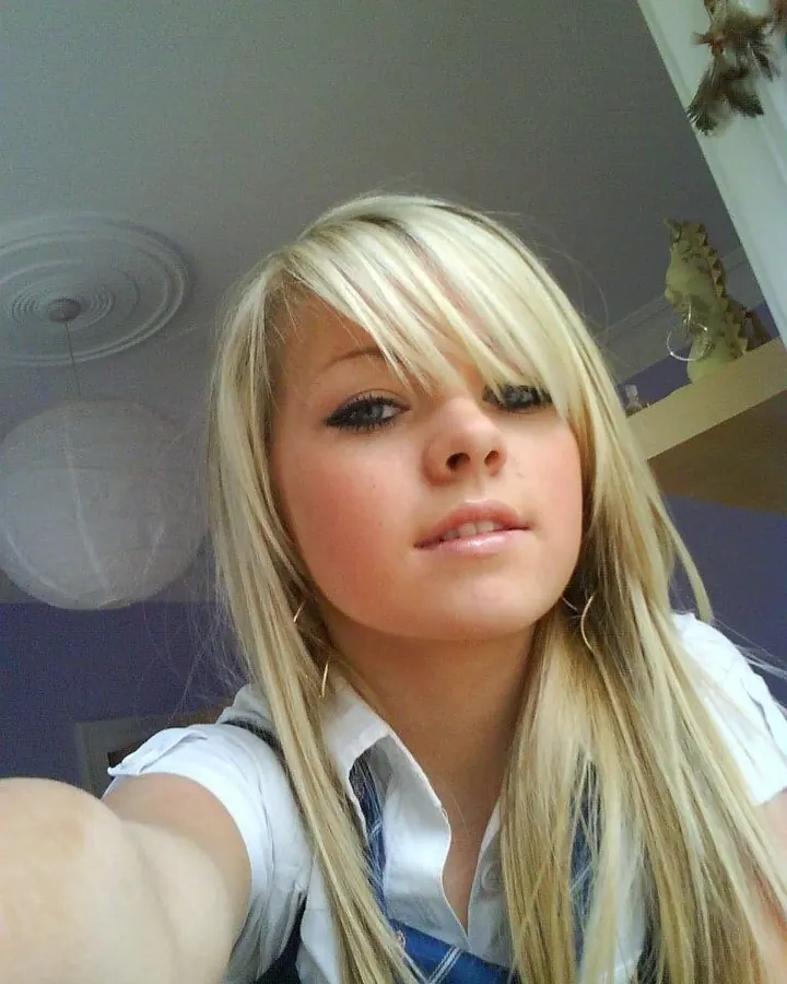 Femme blonde en haut noir et jupe blanche, assise sur une chaise, tenant un téléphone noir et faisant un geste de téléphone.