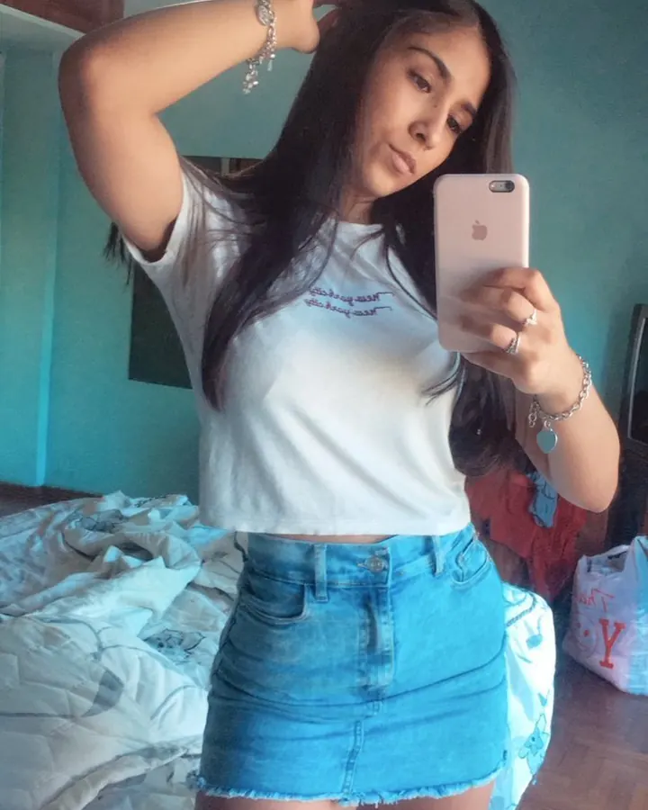 Jeune femme aux cheveux bruns noirs en t-shirt blanc et jupe en jean bleu, prenant un selfie devant un lit défaite.