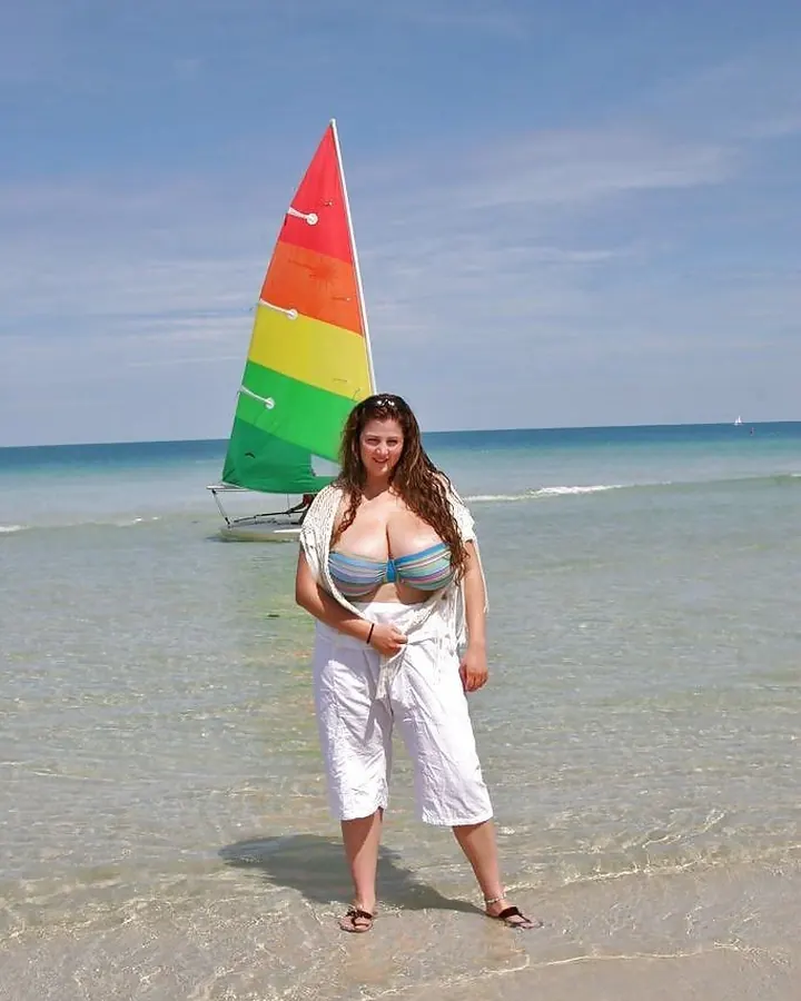 Femme aux cheveux bruns en bikini multicolore, debout dans l'eau claire, avec un voilier coloré en arrière-plan.