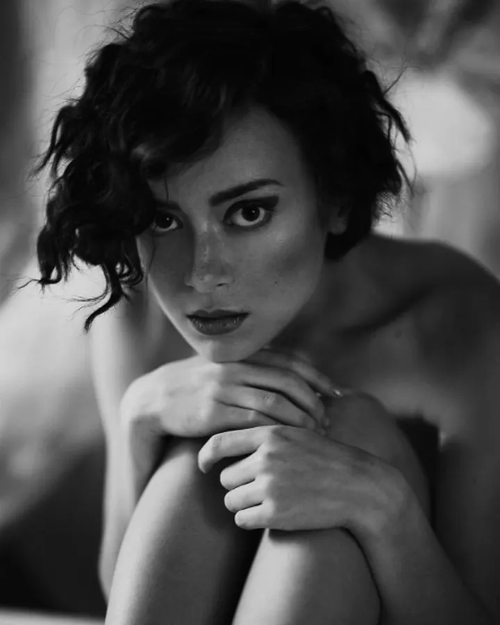 Jeune femme aux cheveux bruns-noirs, portant un haut noir et un short en jean, prenant un selfie devant un miroir dans une chambre moderne.
