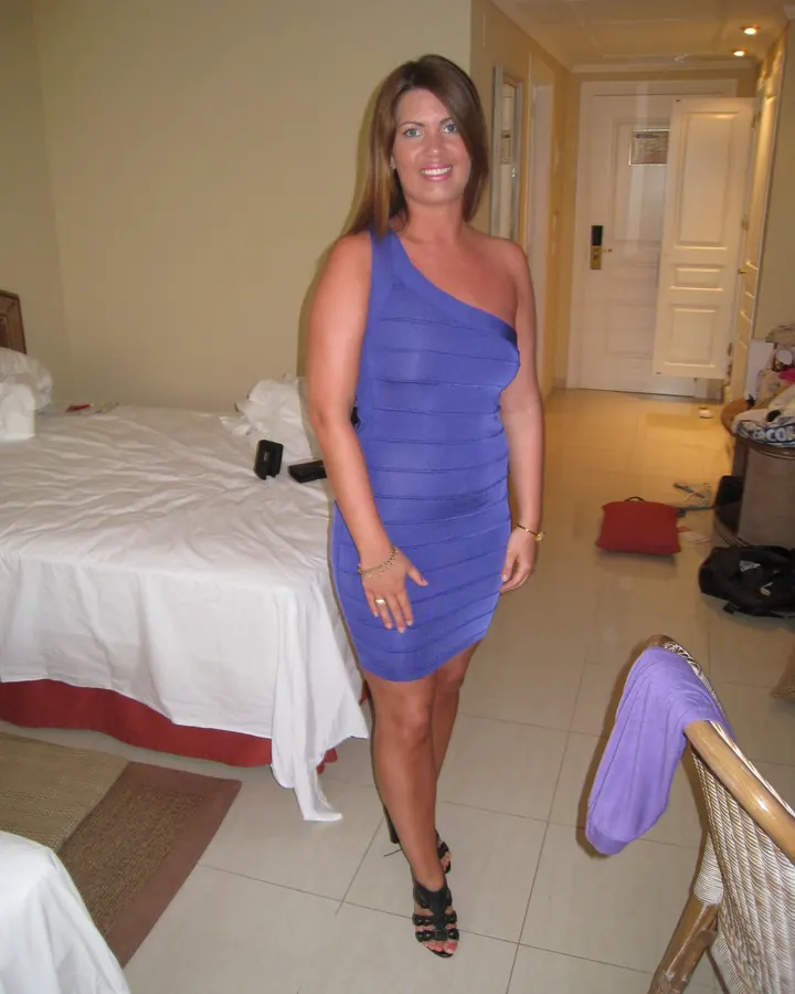 Femme aux cheveux bruns en robe bleue asymétrique, souriant à la caméra dans une chambre d’hôtel.