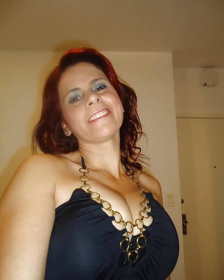 Femme aux cheveux roux en robe noire avec collier en chaînes dorées, souriant à la caméra dans une pièce intérieure.