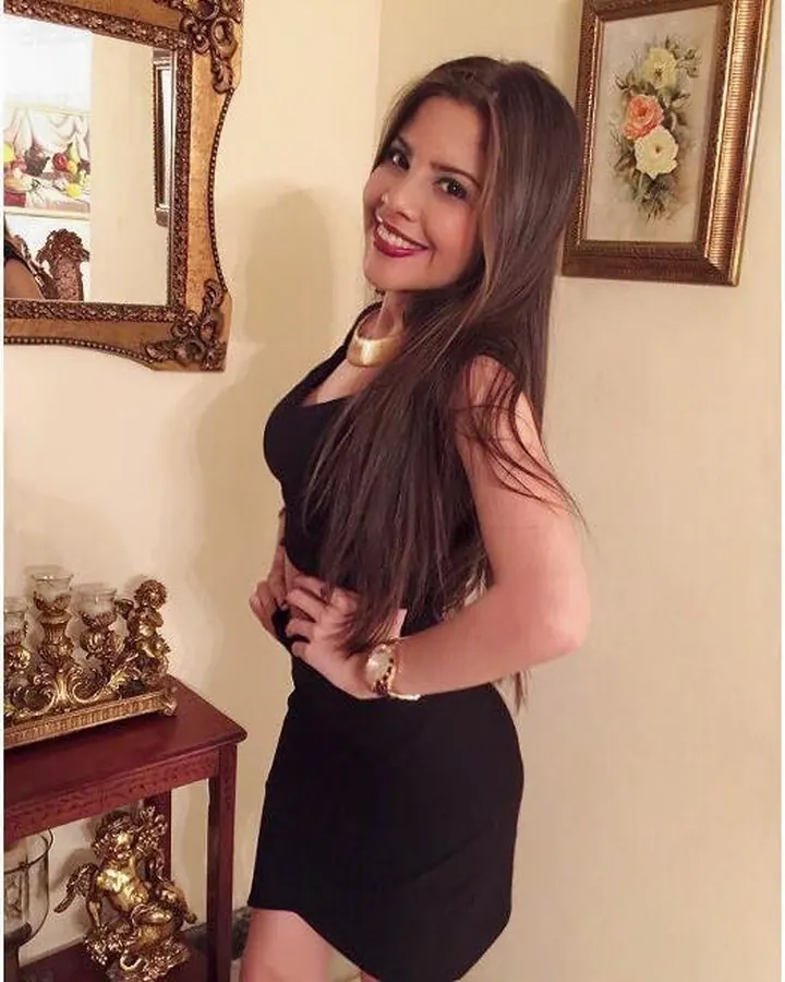 Femme aux cheveux bruns en haut blanc, posant dans une chambre, regardant la caméra.