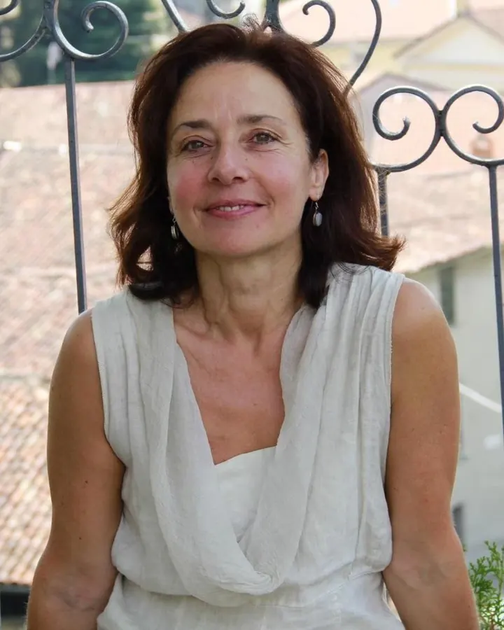 Femme aux cheveux châtains, souriante, assise sur une chaise métallique, devant un fond de clôture en fer forgé.