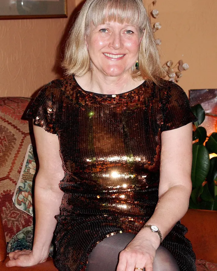 Femme aux cheveux blonds platine, assise sur un canapé, portant une robe scintillante marron et des collants noirs.