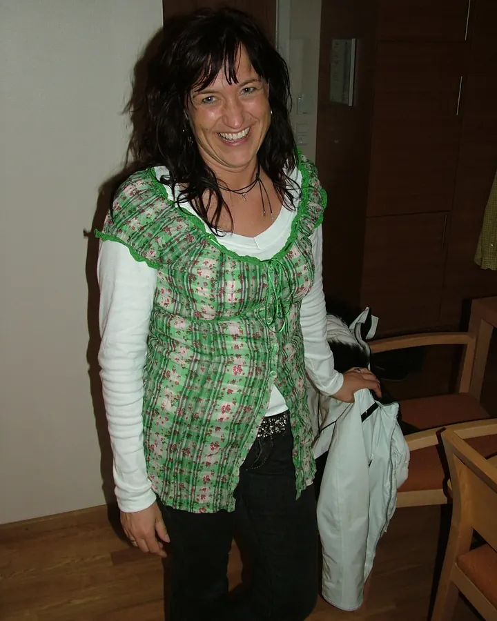 Femme aux cheveux bruns, souriante, portant un haut vert à motifs floraux, debout dans une pièce.