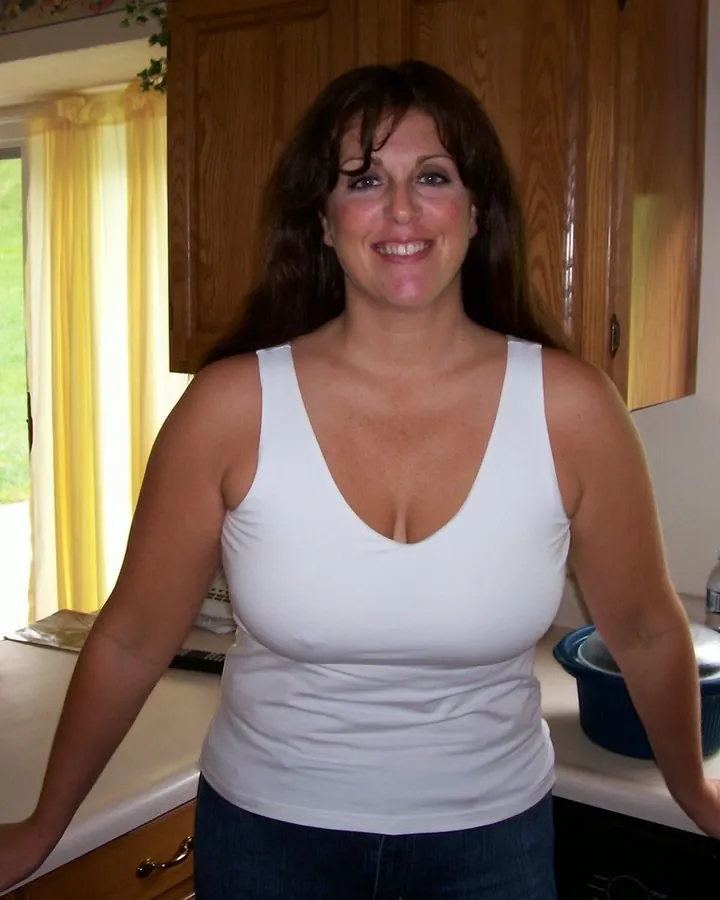 Femme aux cheveux bruns, souriante, debout dans une cuisine, portant un débardeur blanc et un jean.