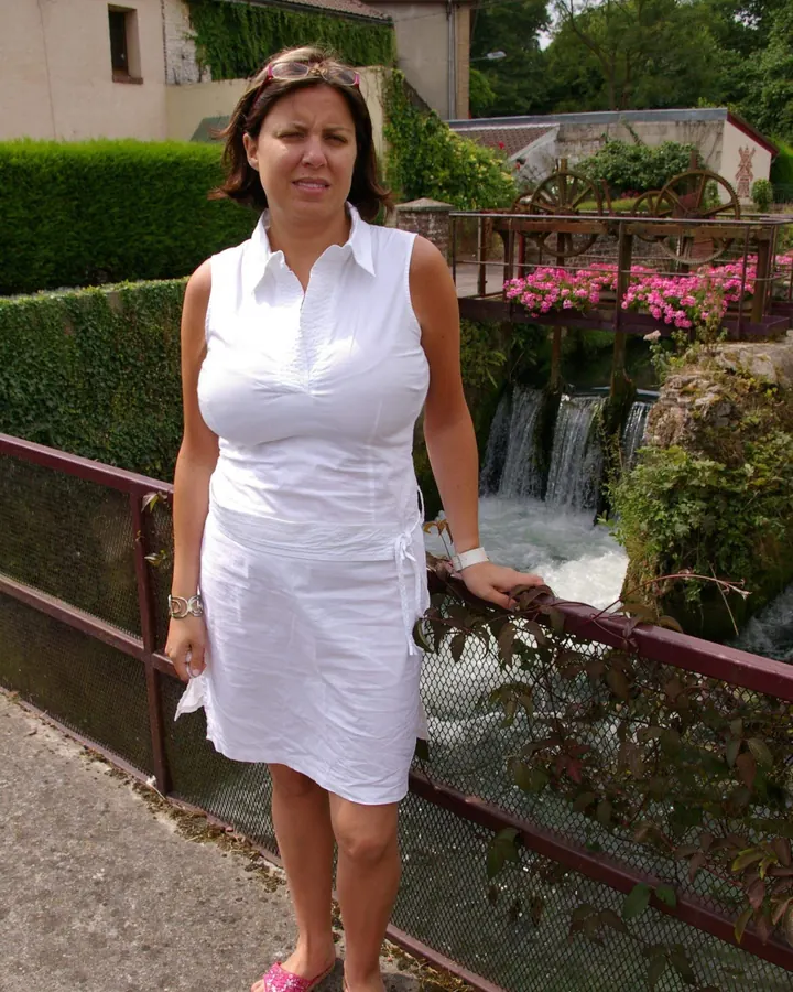 Femme aux cheveux châtains, robe blanche, pose debout près d’un mur blanc, main sur la hanche.