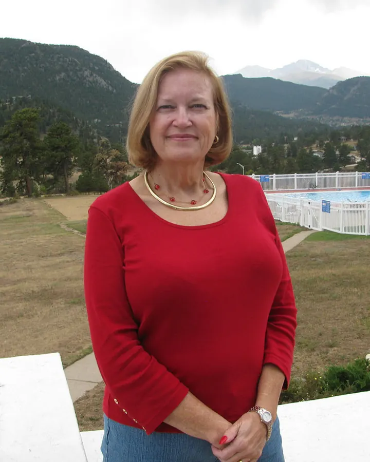 Femme aux cheveux blonds courts, robe rouge, sourire discret, debout devant une terrasse avec vue sur des montagnes.