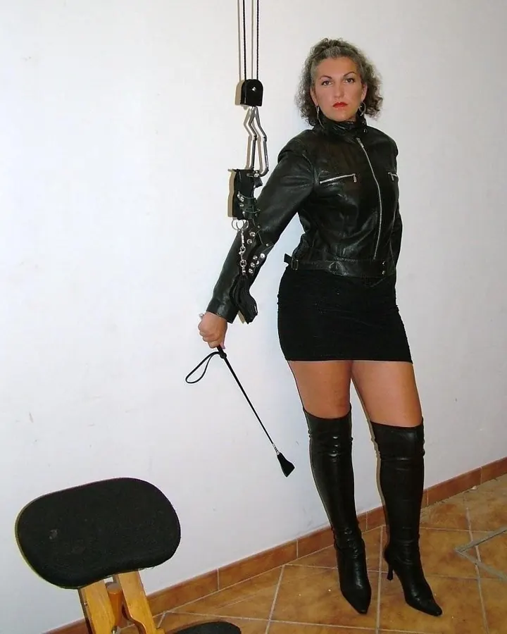 Femme aux cheveux gris, veste en cuir noir, mini-jupe noire, bottes hautes, tenant une cravache, près d’un tabouret.