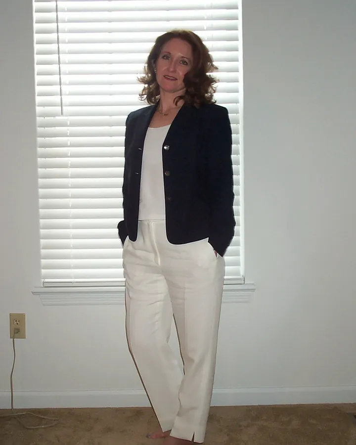 Femme aux cheveux châtains portant un blazer noir et un pantalon blanc, debout devant une fenêtre avec volets blancs, pieds nus sur un tapis beige.