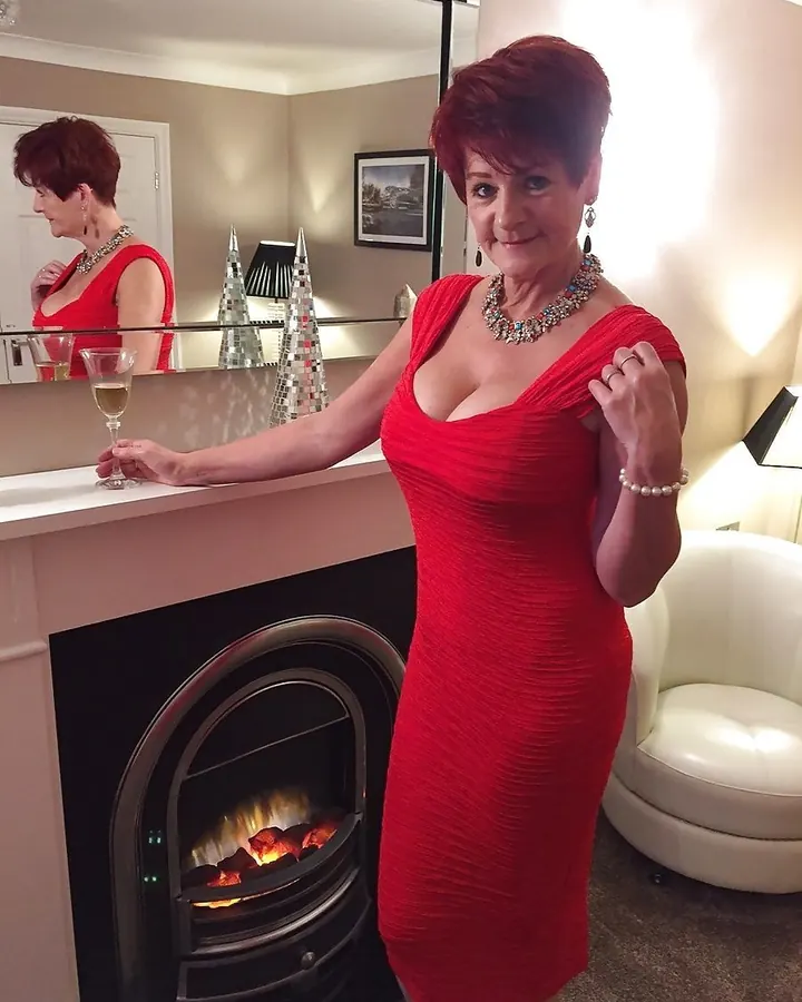 Femme aux cheveux roux portant une robe rouge ajustée, tenant un verre de vin blanc, devant une cheminée allumée, dans une pièce élégante.