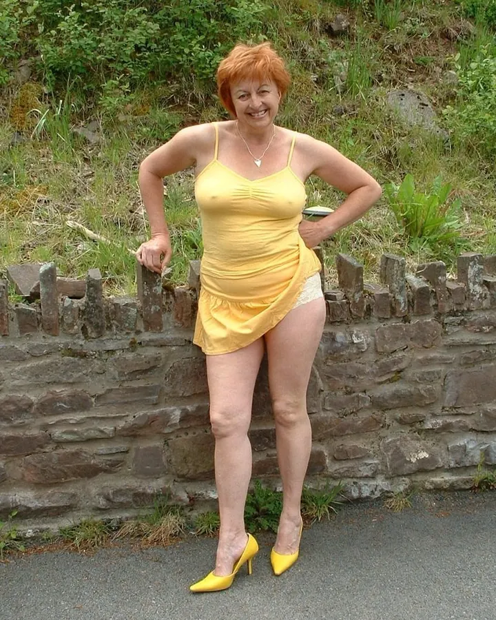Femme aux cheveux roux portant une robe jaune, appuyée contre un mur en pierre dans un cadre naturel.