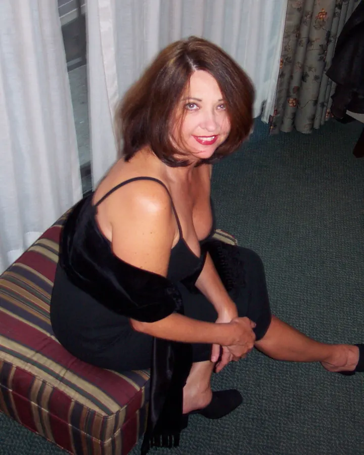 Femme aux cheveux bruns portant une robe noire, assise sur un pouf rayé dans une pièce.