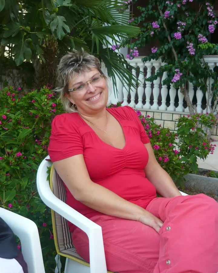 Femme en tenue rouge, assise sur une chaise blanche, entourée de plantes et de fleurs