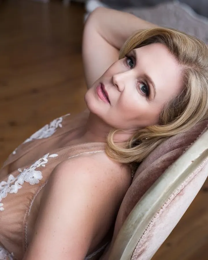 Femme blonde en lingerie beige transparente avec dentelle blanche, allongée sur un fauteuil