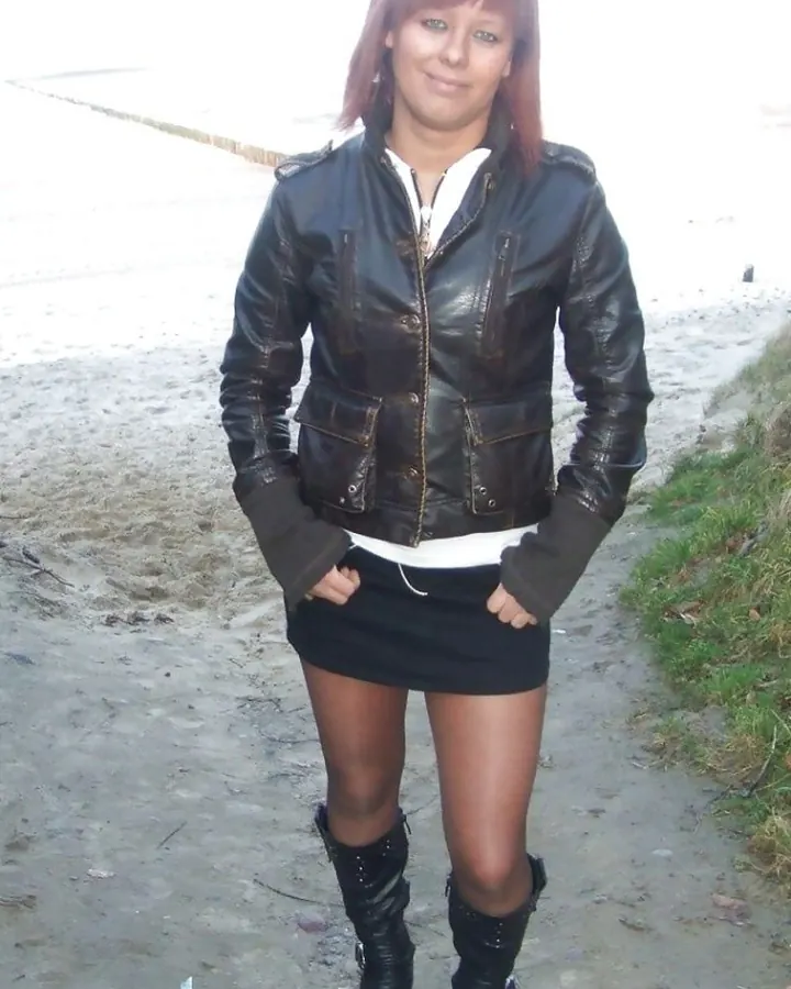 Femme aux cheveux roux portant une veste noire et un haut blanc à mailles, posant devant un mur clair.