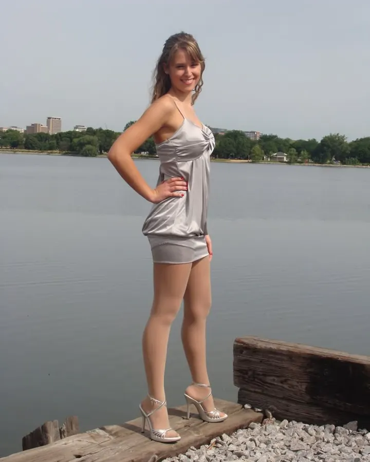 Femme aux cheveux châtains portant un haut noir et une jupe noire, assise sur un banc orné, avec des chaussures à talons noirs.