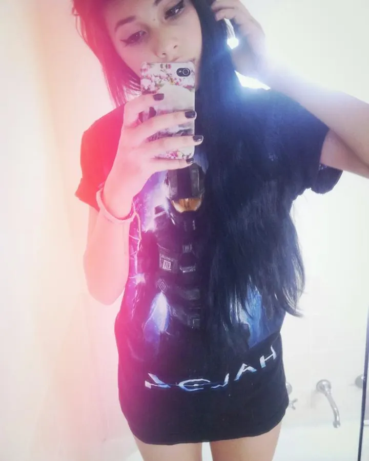 Femme aux cheveux bruns-noirs portant un haut noir et un short noir, prenant un selfie dans une salle de bain blanche.