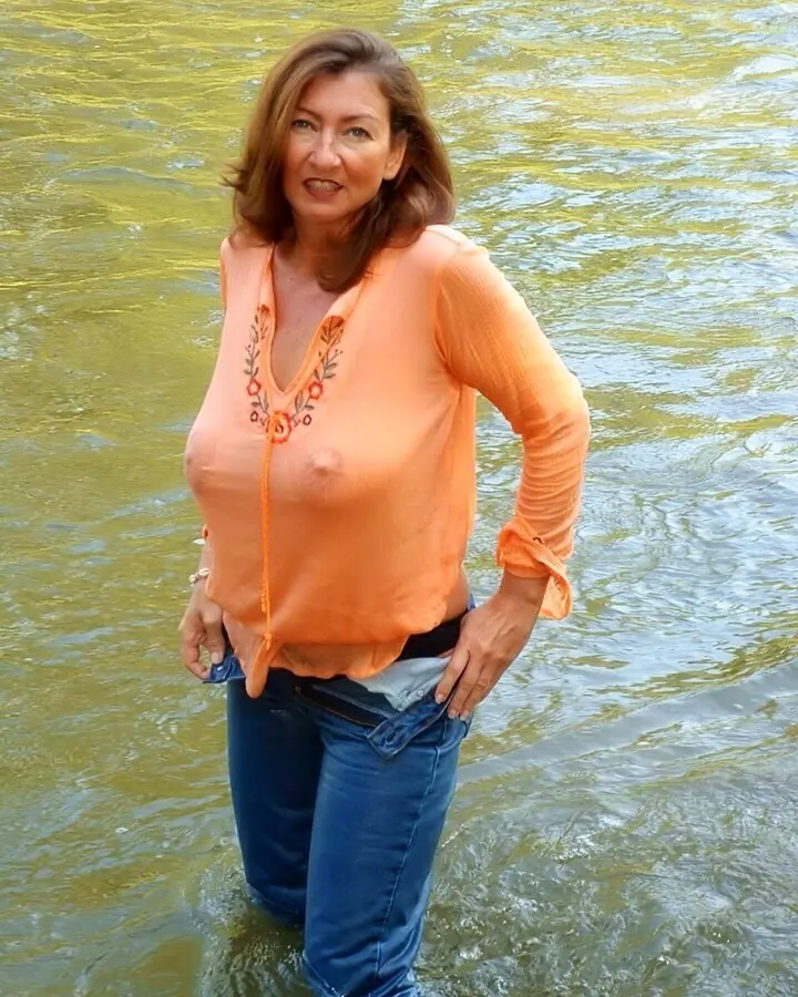 Femme souriante en chemise orange, debout dans l'eau, mains sur les hanches.