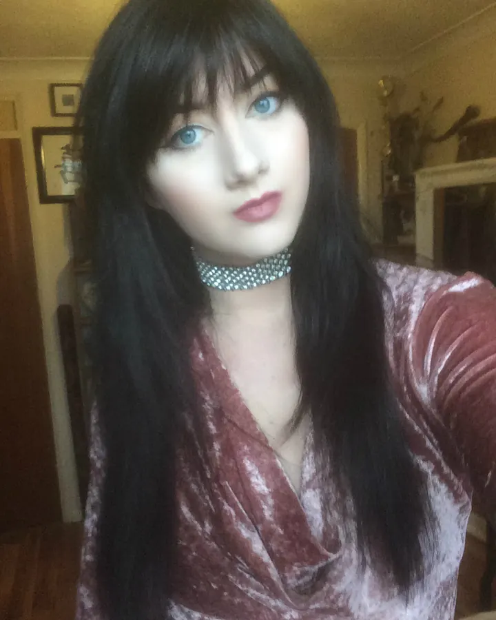 Femme aux cheveux bruns foncés, vêtue d’un haut noir et d’une jupe noire courte, posant dans une pièce avec un canapé beige.