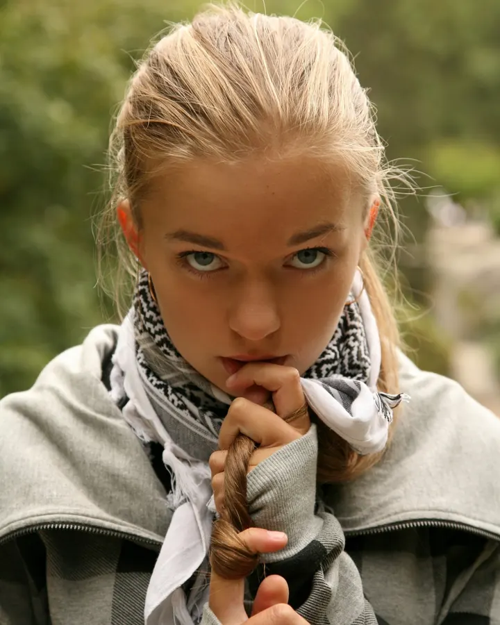Jeune femme aux cheveux blonds, vêtue d’un sweat gris à capuche et d’un foulard, regardant l’appareil photo avec les doigts sur les lèvres.