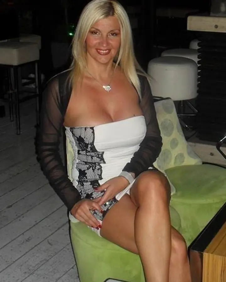 Femme aux cheveux blonds, souriante, portant une robe noire, assise sur un canapé, dans une chambre.