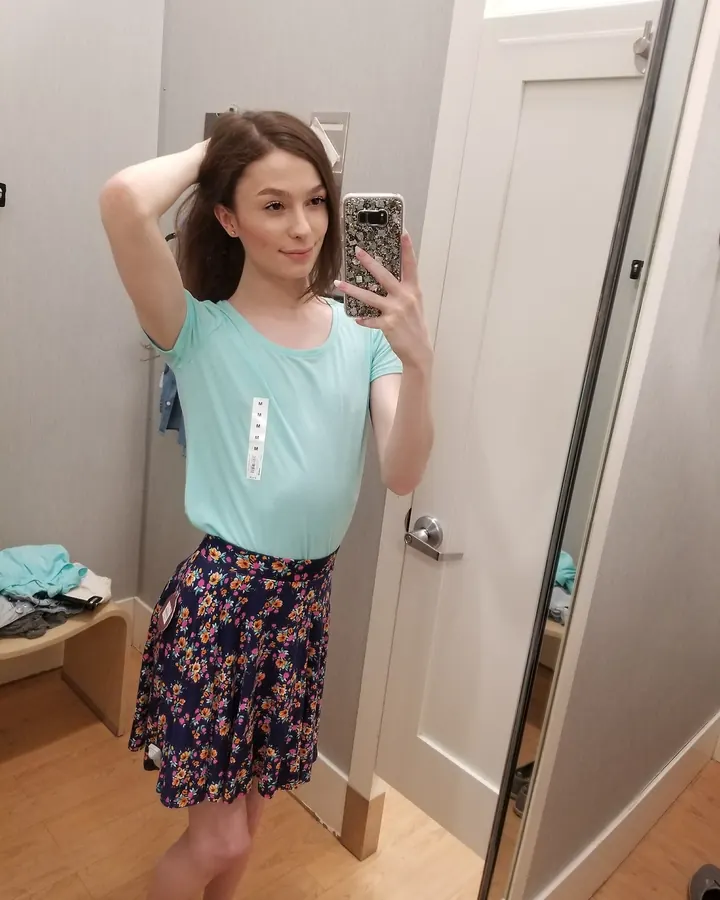 Jeune femme européenne aux cheveux châtains, prenant un selfie devant un miroir dans une cabine d’essayage, portant un haut turquoise et une jupe fleurie.