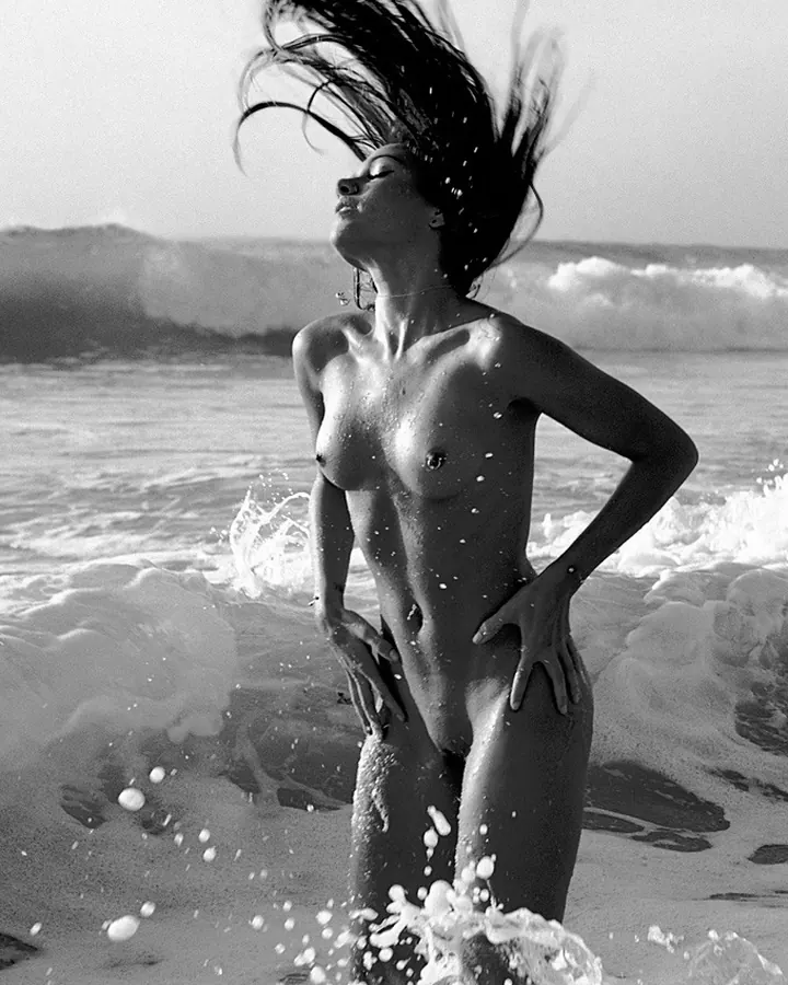 Jeune femme européenne aux cheveux bruns-noirs, portant un haut noir décolleté, posant dans une cuisine, souriant légèrement.