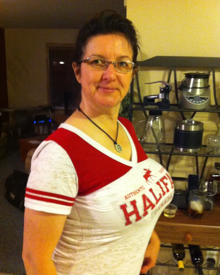 Femme aux cheveux bruns-noirs portant des lunettes et un t-shirt blanc et rouge avec l'inscription "HALIFE", souriante devant une étagère avec des bouteilles et des verres.