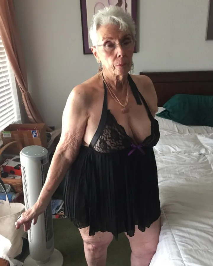 Femme aux cheveux blancs en tenue noire, debout dans une chambre, regardant l'appareil.