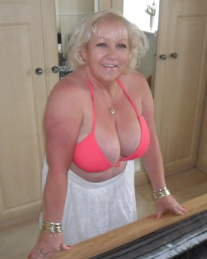 Femme blonde en robe noire, souriante, les mains sur les hanches, devant un rideau.