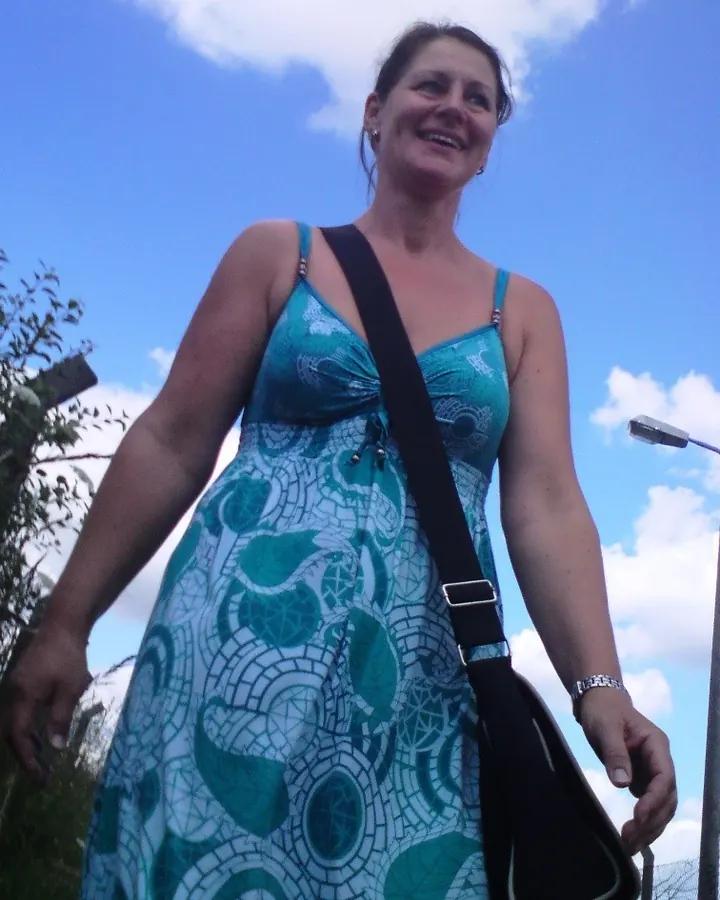 Femme souriante en robe turquoise à motifs, portant un sac noir, sous un ciel bleu.