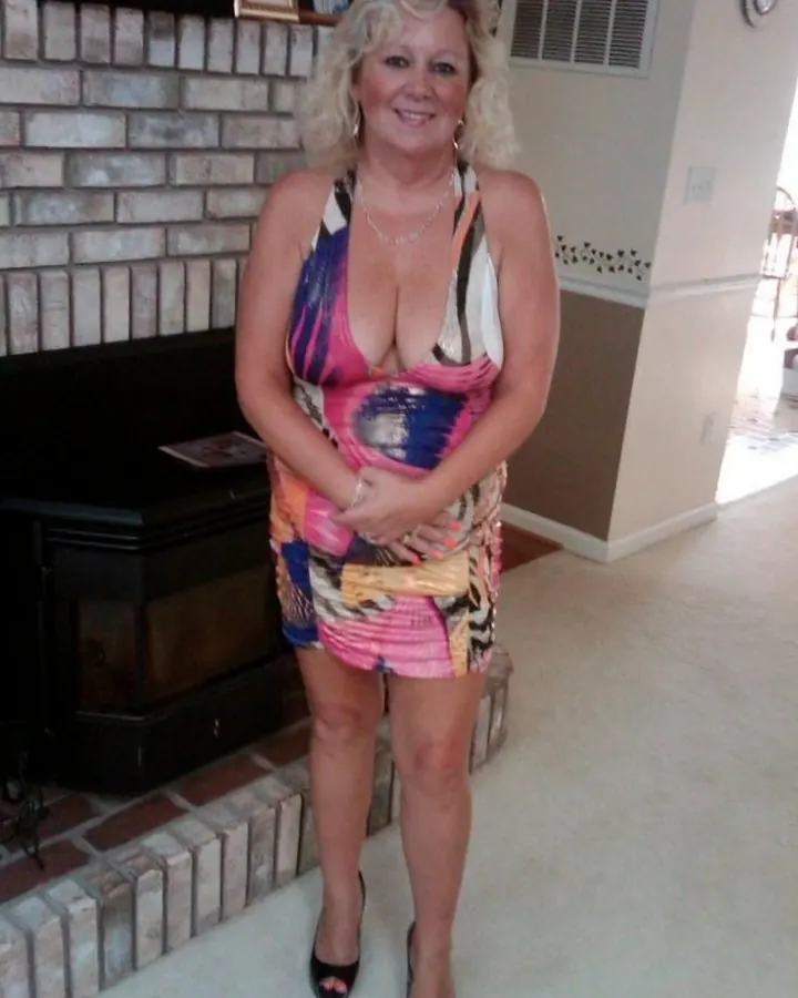 Femme aux cheveux blonds platine, souriante, portant une robe multicolore, assise sur un canapé beige, pieds en sandales vertes.