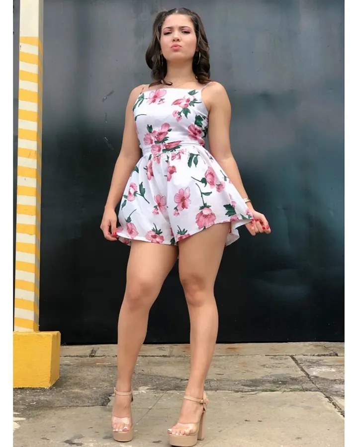 Femme aux cheveux bruns foncés, portant une robe fleurie blanche et rose, posant de face contre un mur noir, les pieds en sandales compensées.