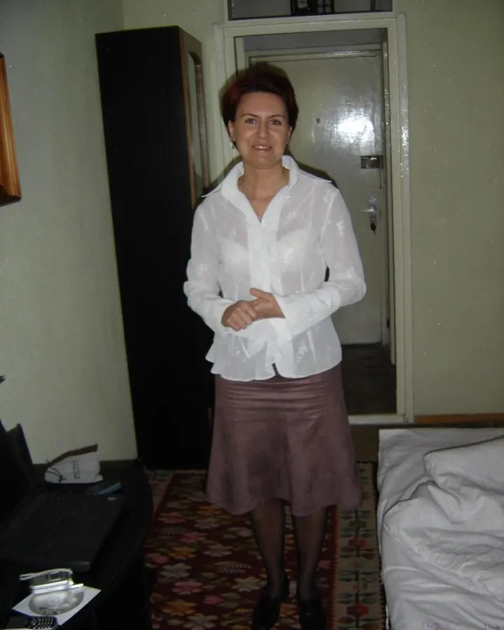 Femme aux cheveux courts roux en chemise blanche transparente et jupe marron, debout dans une chambre.