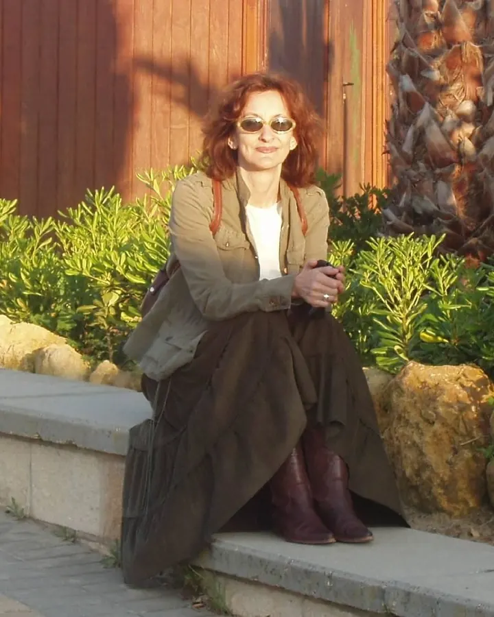 Femme aux cheveux châtains, portant une veste verte, une robe noire et des bottes marron, assise sur un mur extérieur près d’une palme.