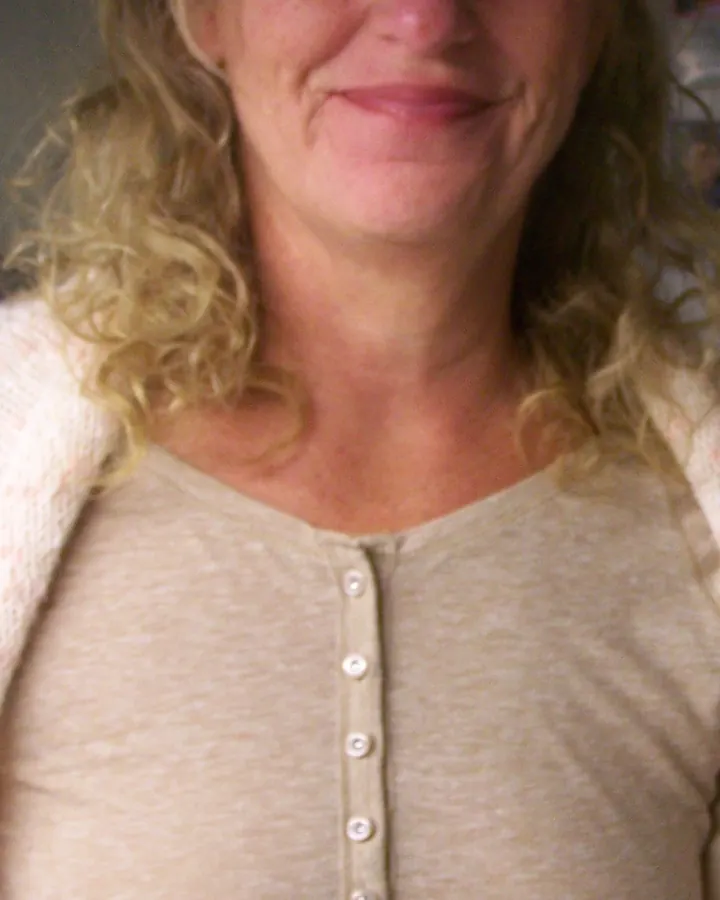 Visage souriant d'une femme blonde aux cheveux bouclés, portant un haut beige boutonné, dans un cadre intérieur doux.