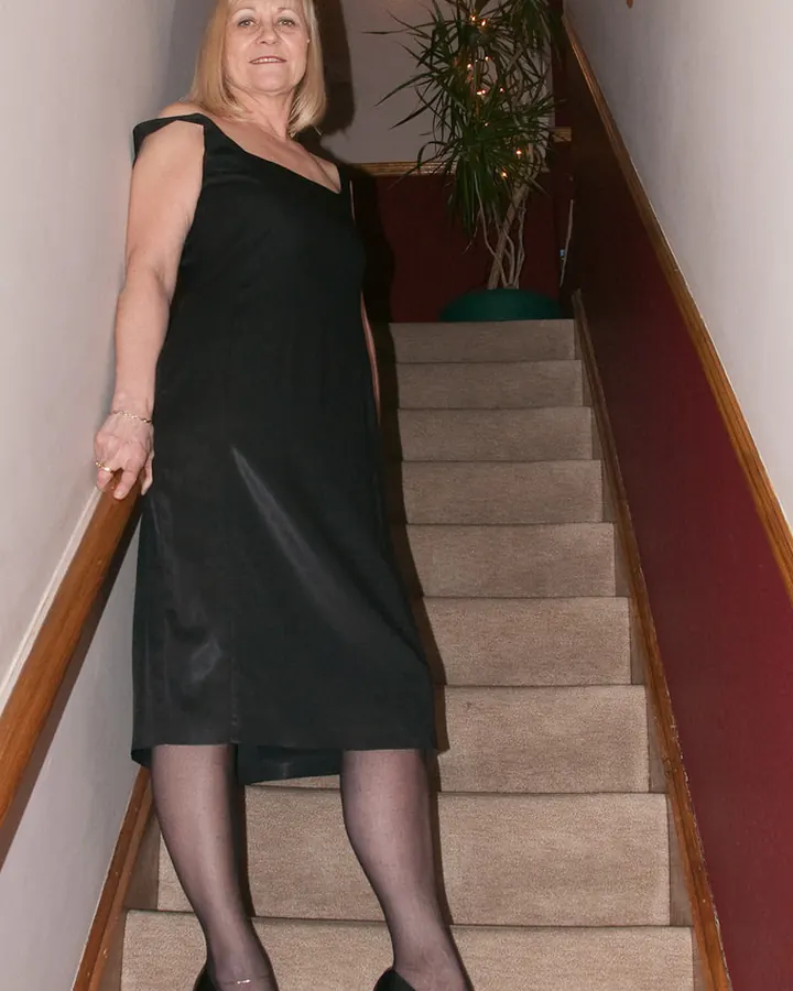 Femme aux cheveux blonds portant une robe noire, debout sur un escalier en moquette beige avec un mur rouge et une plante verte en arrière-plan.