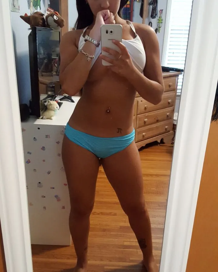 Jeune femme aux cheveux bruns-noirs portant un haut blanc et un short turquoise, prenant un selfie devant un miroir dans une chambre avec un bureau blanc.