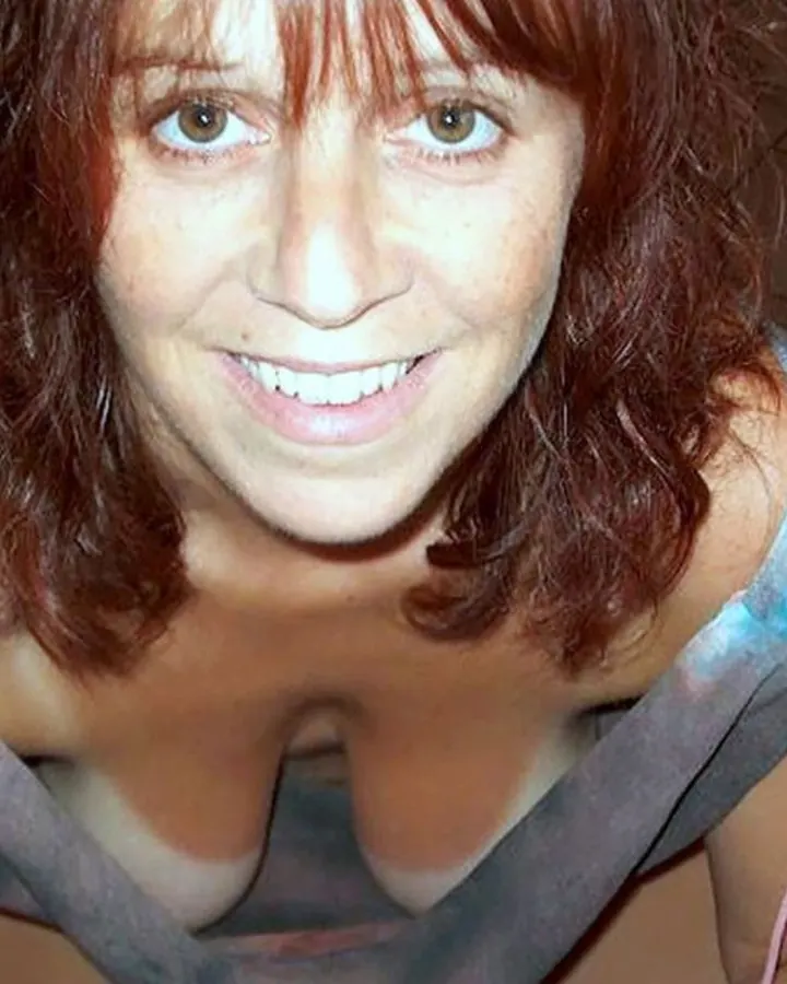 Femme aux cheveux roux portant un haut transparent bleu, souriante en position accroupie sur un sol carrelé marron.