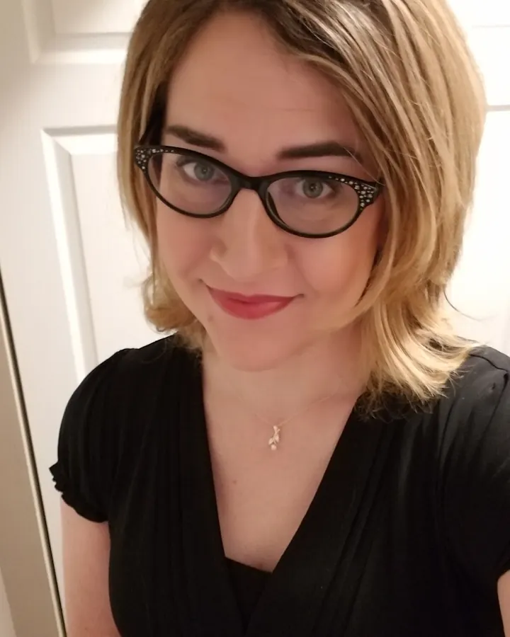 Femme aux cheveux blonds portant un haut gris sans bretelles, assise sur une table blanche dans un restaurant extérieur avec des chaises noires.