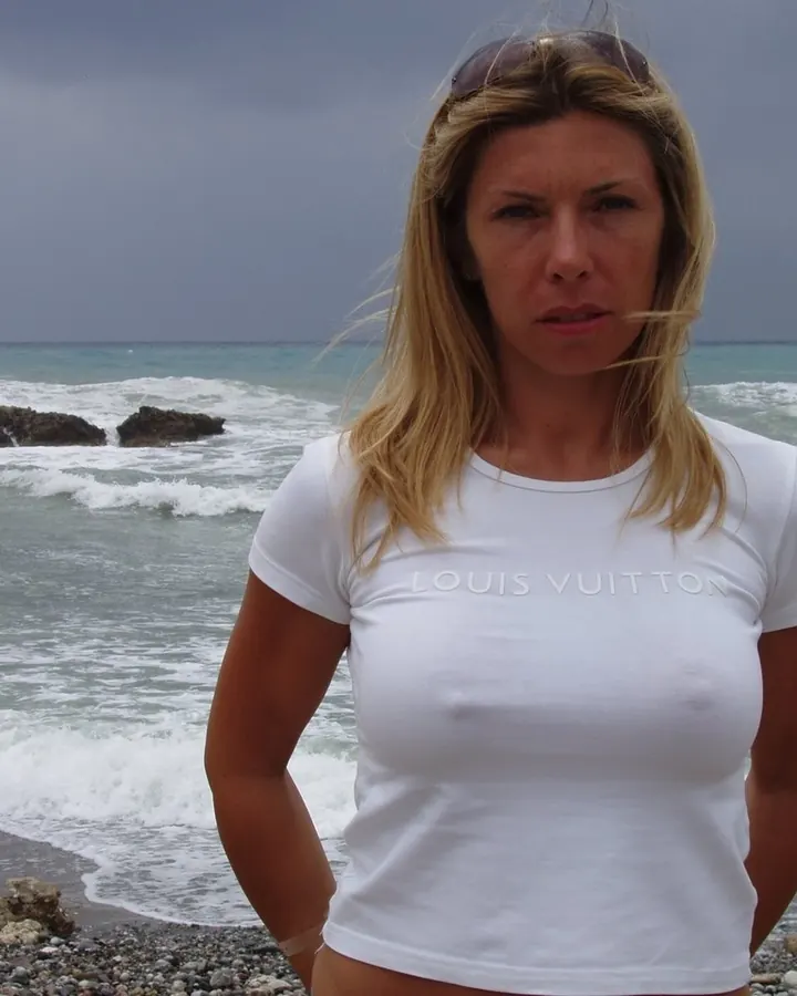 Femme blonde en t-shirt blanc "LOUIS VUITTON", debout sur une plage rocheuse, regardant l'appareil avec une expression intense.