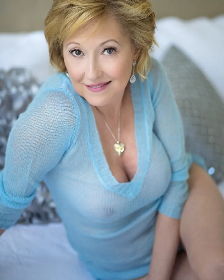 Femme aux cheveux blonds, souriante largement, debout devant un mur blanc, regardant l'appareil avec une expression amicale.