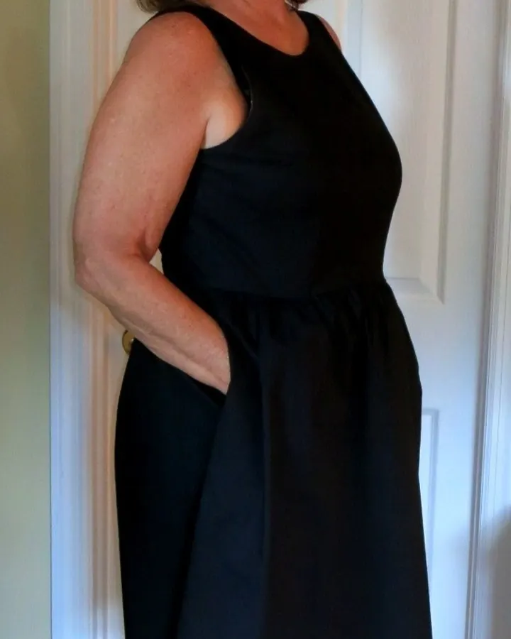 Femme aux cheveux châtains, portant une robe noire sans manches, souriante, debout devant une porte blanche, chaussée de sandales à talons.